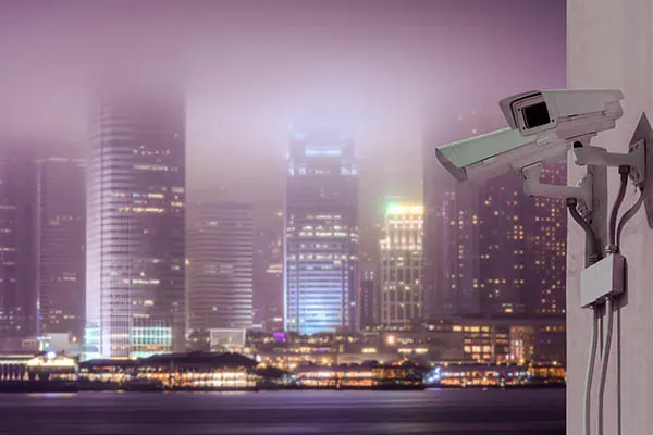 Two white security cameras on a wall with a blurry, hazy city skyline and water in the background at night