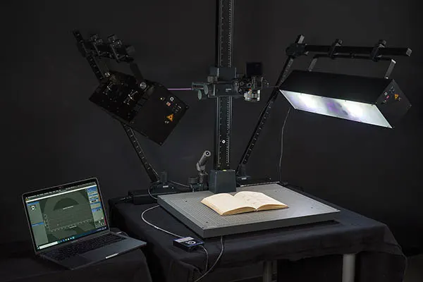 An open book on a scanning platform with adjustable lights and a camera above, with a laptop on the left for standardized camera testing