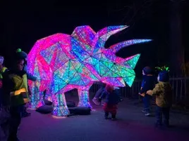 Children stand by a large, colorful, illuminated triceratops sculpture at night, captured without a camera luminance meter