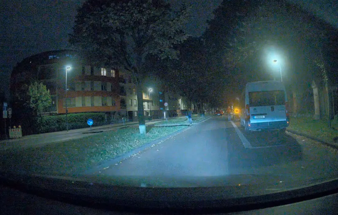 夜间一条昏暗街道的低分辨率监控摄像头画面,画面中可见一栋公寓楼、树木、路灯以及一辆停放的厢式货车