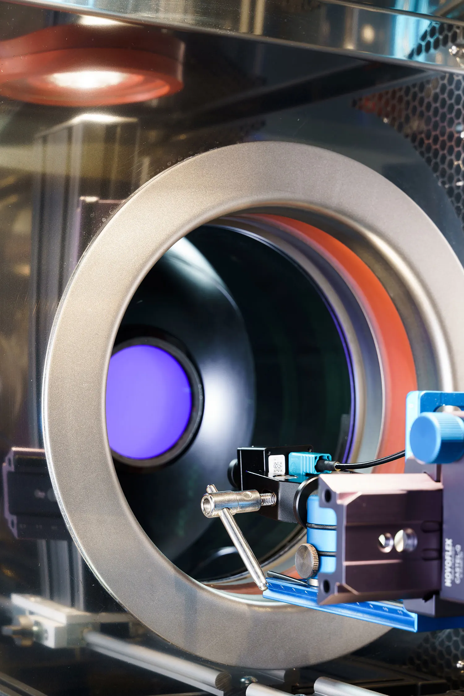Close-up of a large circular opening in a machine, with a blue glowing light at its center and complex equipment for automotive camera im...