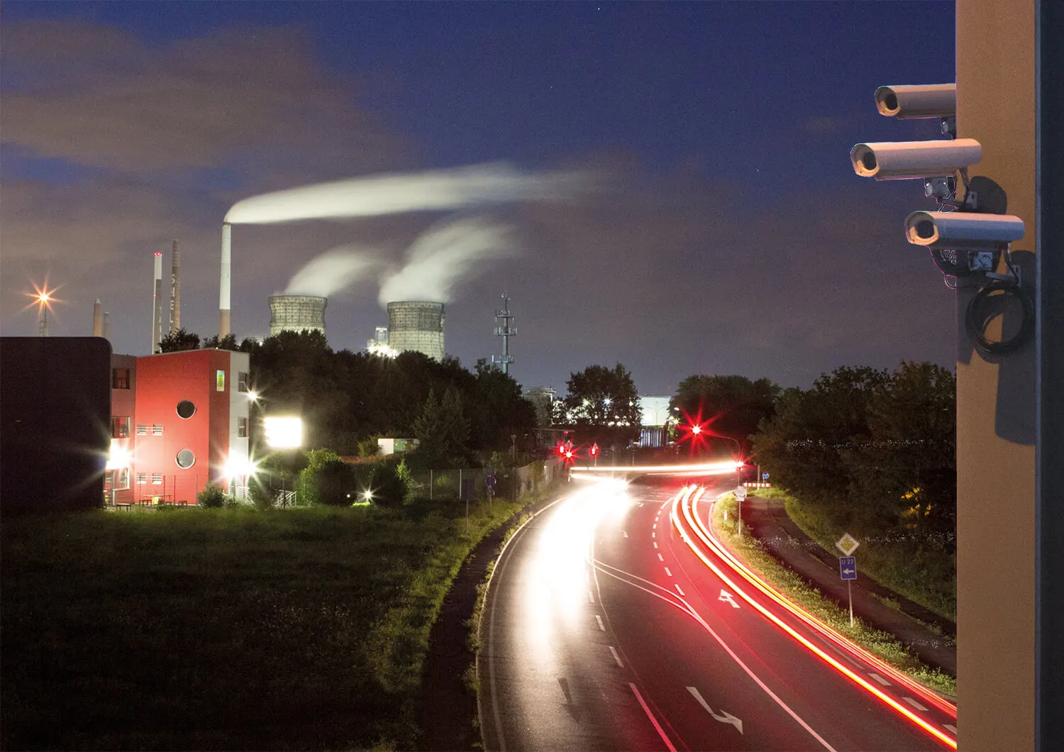 高速公路的夜景,可见车灯留下的光轨,冒着烟的发电厂,一栋红色建筑以及三台监控摄像头