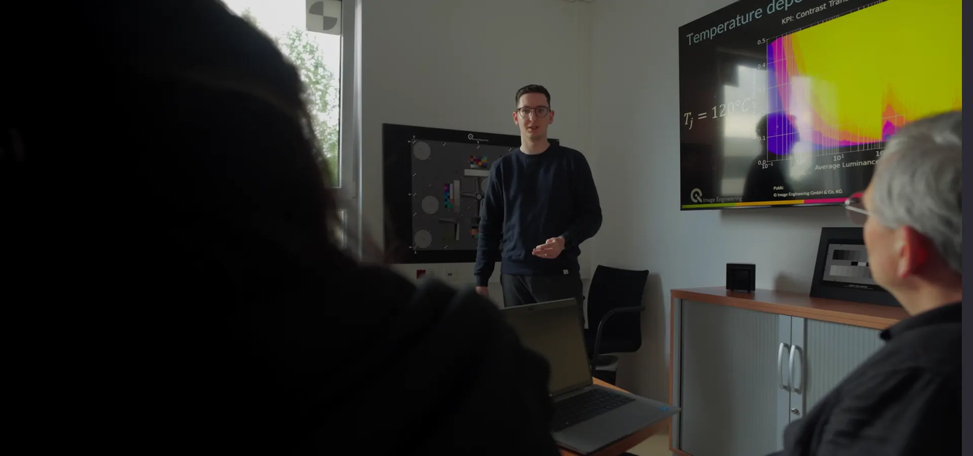 A man presents a data visualization on a large screen to two attendees, illustrating an image quality concept