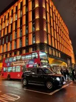A double-decker bus, black taxi, and pedestrians on a street at night, with a camera luminance meter on a building facade