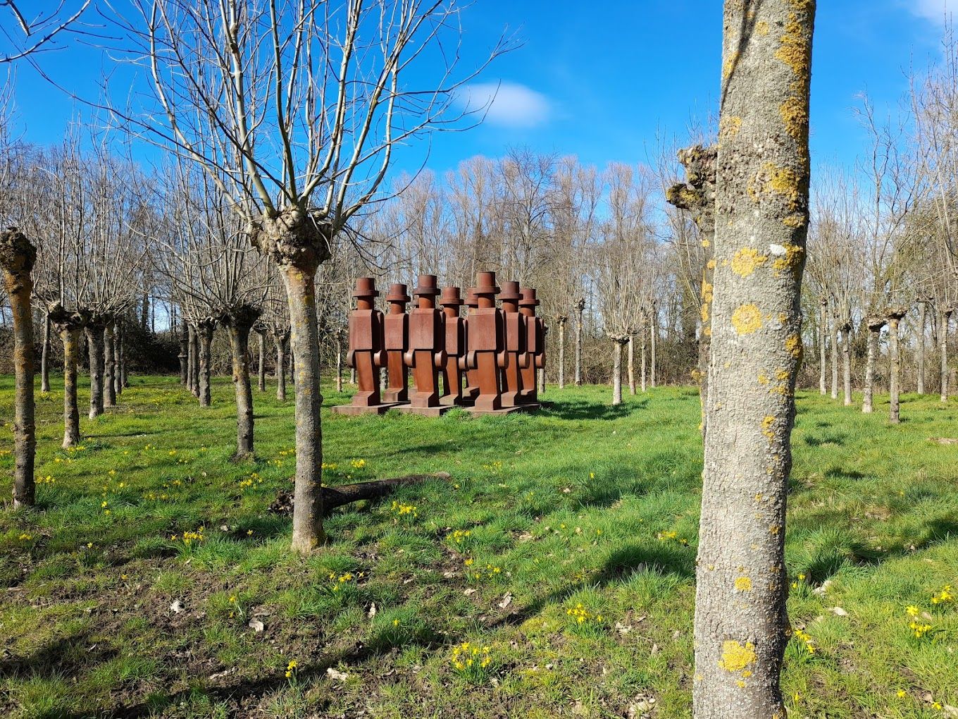 Pollarded trees and green grass with yellow flowers surround a row of weathered metal figures under a blue sky