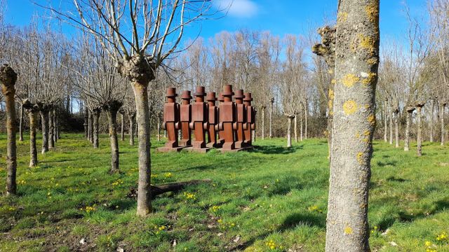 Pollarded trees and green grass with yellow flowers surround a row of weathered metal figures under a blue sky